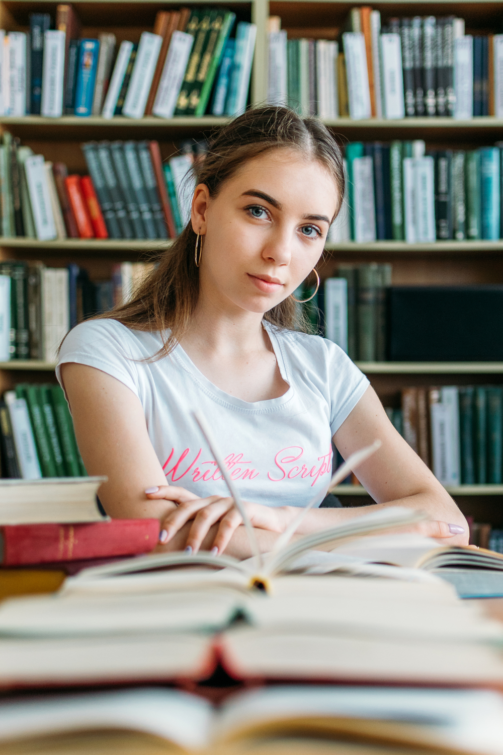 Ung kvinna sitter vid ett bord med böcker i ett bibliotek och tittar mot kameran.