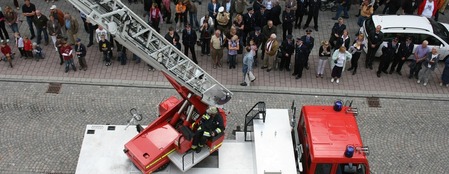 Vy ovanifrån av en brandbil med stege och brandmän, omgiven av en folksamling på en gata.