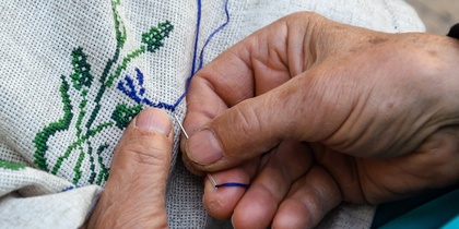 Närbild av händer som broderar med blå och gröna trådar på vitt tyg.