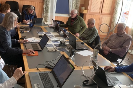 En grupp vuxna sitter runt ett konferensbord med bärbara datorer i ett ljust mötesrum.