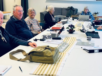 En grupp äldre personer sitter runt ett konferensbord i ett ljust mötesrum med datorer och anteckningar.