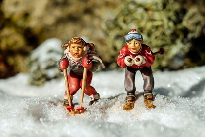 Två små figurer på skidor står i snö, klädda i vinterkläder och ser ut att åka utför.