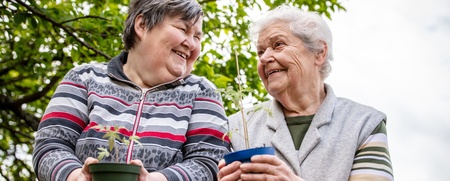 Två äldre kvinnor står utomhus och håller varsin blomkruka, de ler mot varandra.