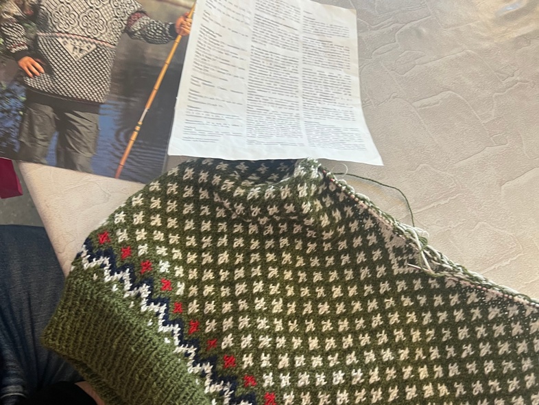 En person stickar en grön tröja med mönster, bredvid ett mönsterblad och en bild på en färdig tröja.