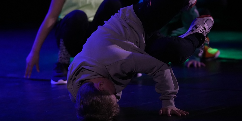 En person utför ett breakdance-move på golvet med stöd av ena handen, andra personer i bakgrunden.