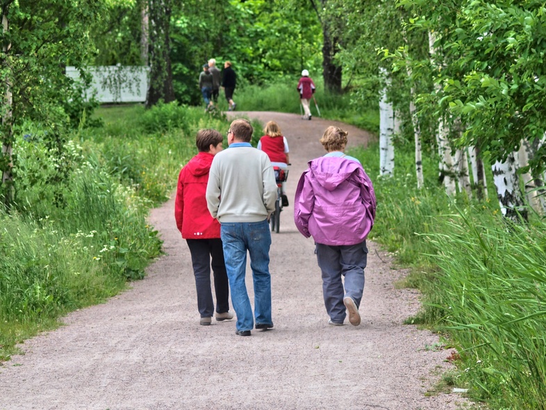 Flera personer promenerar på en grusväg omgiven av gröna träd och gräs en sommardag.