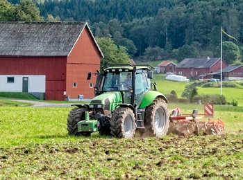 En grön traktor plöjer en åker framför en röd ladugård på landsbygden med skog i bakgrunden.