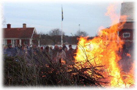 En grupp människor står utomhus och tittar på en stor brasa, med svenska flaggan i bakgrunden.