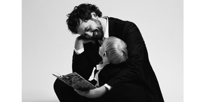En man i kostym sitter barfota på golvet och läser en bok för ett litet barn, båda ser glada ut.