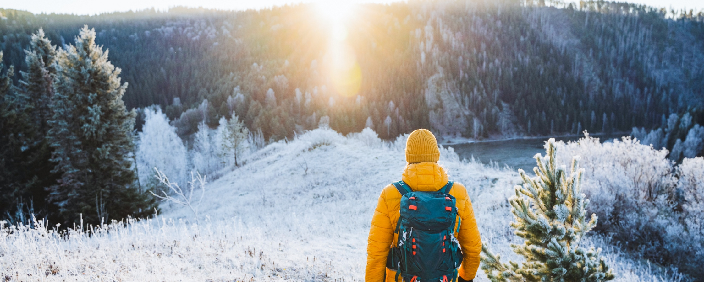 Person med gul jacka och ryggsäck står i ett frostigt vinterlandskap och ser mot solen över en skog.