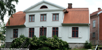 Gammalt vitt trähus med rött tegeltak och röda fönster, omgiven av buskar och träd.