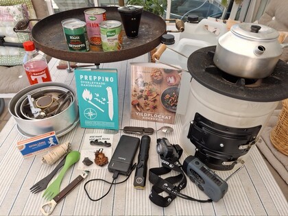 Utrustning för prepping och överlevnad på ett bord, inklusive mat, kokkärl, verktyg och böcker.