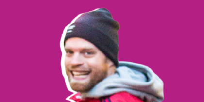 Man i röd jacka och svart mössa pekar och ler mot kameran, lila bakgrund.