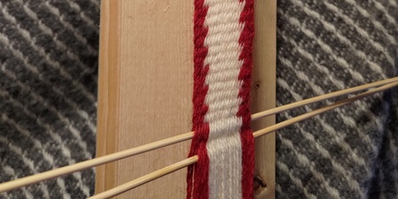 Närbild av ett bandvävningsprojekt med rött och vitt garn på en träplatta.