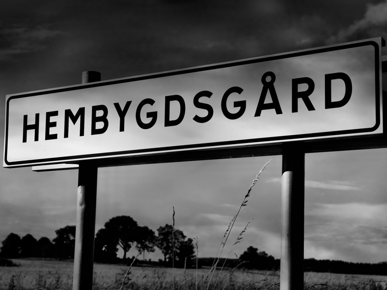 Vägskylt med texten 'HEMBYGDGÅRD' mot en landsbygd med träd och fält under en dramatisk himmel.