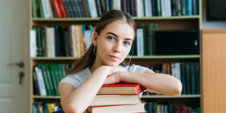 En ung kvinna sitter vid ett bord i ett bibliotek med en hög böcker framför sig.