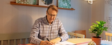 En man med glasögon sitter vid ett köksbord och skriver i ett block. En grön växt står på bordet.