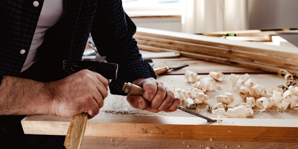 En person arbetar med träbearbetning, använder en mejsel och hammare på en träplanka.