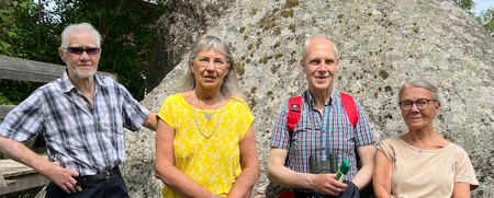 Fyra vuxna personer står framför en stor sten utomhus, med träd och blå himmel i bakgrunden.