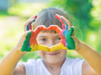 Ett barn med färgglada målade händer formar ett hjärta framför sitt ansikte utomhus.