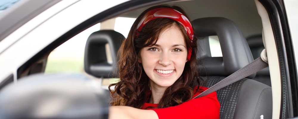 Ung kvinna med långt brunt hår och röd tröja sitter leende bakom ratten i en bil.