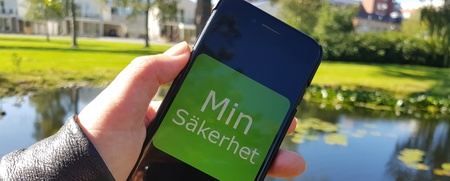 En hand håller en mobiltelefon med texten 'Min Säkerhet' framför en damm i en park.