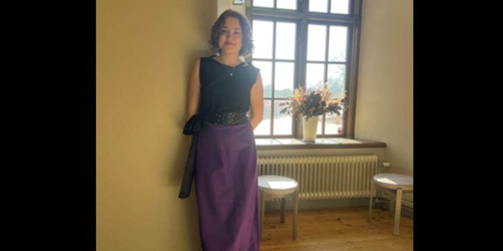 Person i svart topp och lång lila kjol står mot en vägg i ett solbelyst rum med trägolv och stora fönster.