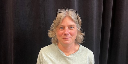 En person med ljust hår och glasögon på huvudet står framför ett mörkt draperi och tittar mot kameran.