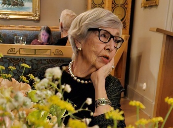 En kvinna med pärlhalsband sitter vid ett bord med blommor i ett elegant rum med konst på väggarna.