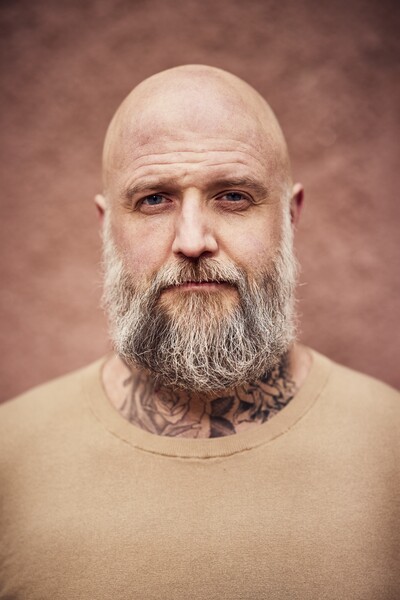 Skäggig, skallig man med tatueringar på halsen, beige tröja och neutral bakgrund.
