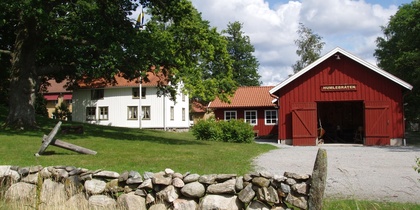 Gårdsmiljö med röd lada, vit byggnad, stengärdsgård och grönskande träd under blå himmel.