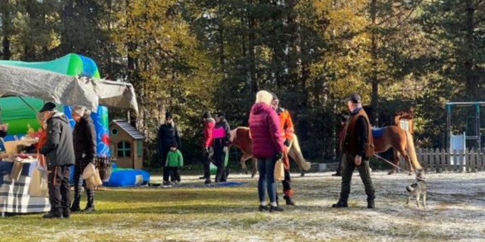 Människor på en utomhusmarknad med hästar, hund och tält, omgiven av skog och lekplats.