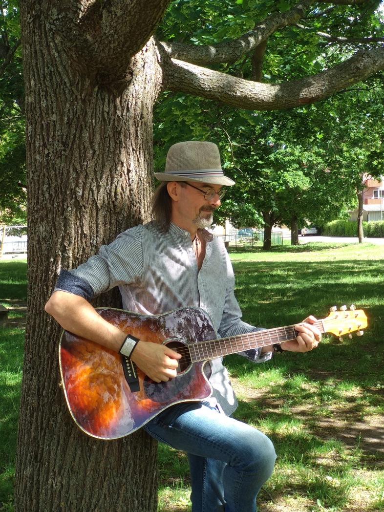 Mathias Beltramo Brandin med hatt spelar akustisk gitarr och lutar sig mot ett träd i en solig park.
