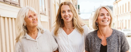 Tre kvinnor går arm i arm på en solig gata med trähus.