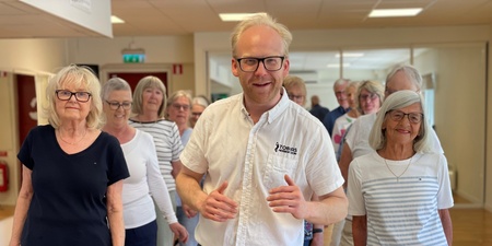 En grupp äldre personer och en yngre man står tillsammans inomhus, leende och avslappnade.