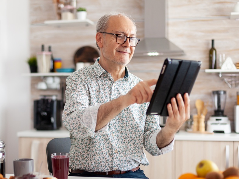 Äldre man sitter vid köksbordet, ler och använder en surfplatta. Frukost med kaffe och frukt på bordet.