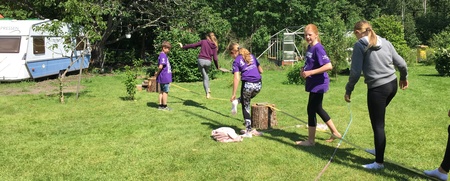 Flera ungdomar går på lina utomhus på en gräsmatta med träd, husvagn och växthus i bakgrunden.