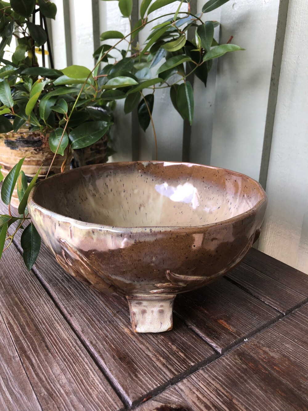 En handgjord keramikskål med brun glasyr står på ett träbord bredvid en grön växt.