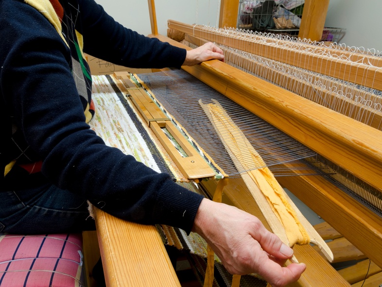 En person väver tyg på en stor vävstol i ett ljust rum. Händerna arbetar med trådarna.