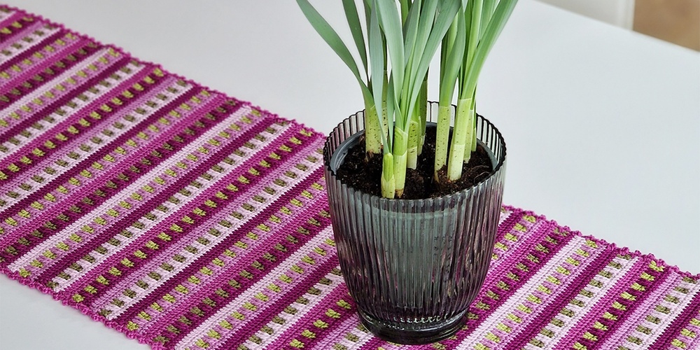 En svart kruka med gula påskliljor står på en vit bordsyta med en färgglad löpare i rosa och lila toner.