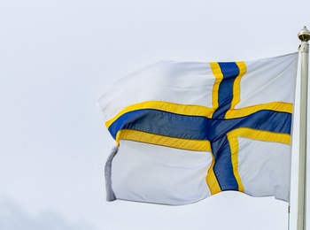 Flagga med blått kors och gul kant på vit bakgrund vajar mot en molnig himmel.