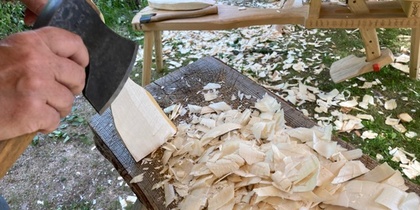 En person täljer trä med en yxa utomhus, träspån ligger på ett bord.