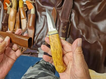 En person håller en handgjord kniv med trähandtag och visar dess läderslida på ett bord med flera andra knivar.