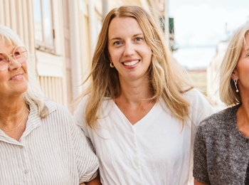 Tre kvinnor går arm i arm på en solig gata med trähus.