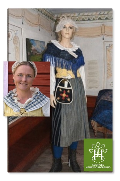 Kvinna i traditionell svensk folkdräkt med förkläde och sjal, inomhus i historisk miljö.