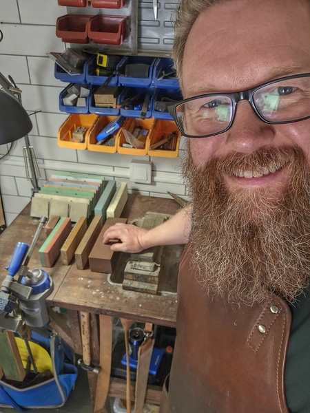 Man med skägg och glasögon står vid arbetsbänk med slipstenar och verktyg i verkstad.