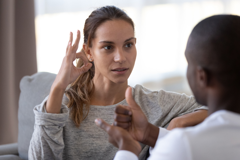 Två personer kommunicerar med teckenspråk i ett ljust vardagsrum.