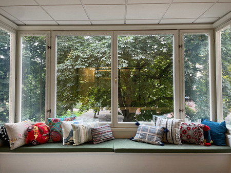 Stora fönster med utsikt mot en park med gröna träd. Kuddar i olika mönster och färger ligger på sittdynor framför fönstren.