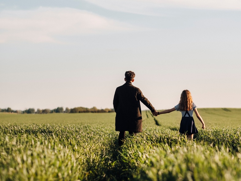 Två personer, en vuxen och ett barn, går hand i hand genom en grön åker under blå himmel.