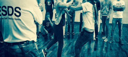 En grupp människor tränar självförsvar i en sal med trägolv, en instruktör syns i förgrunden.